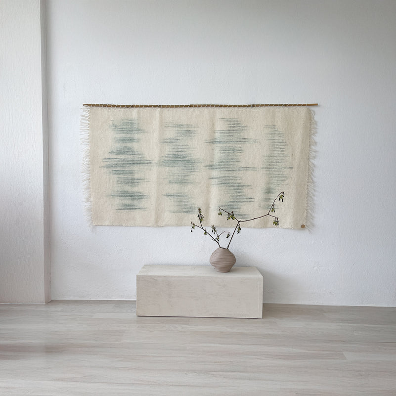 Handwoven Matcha wool tapestry featuring a soothing green olive dip-dyed design on raw white wool, suspended on a bamboo rod, styled in a minimalist interior with a ceramic vase and natural decor elements.
