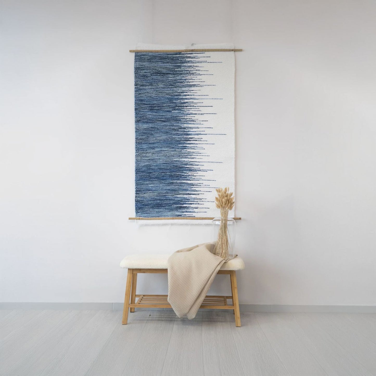 Handwoven white tapestry with a gradient blue zigzag pattern, suspended on a bamboo rod, ideal for modern and minimalist interiors.