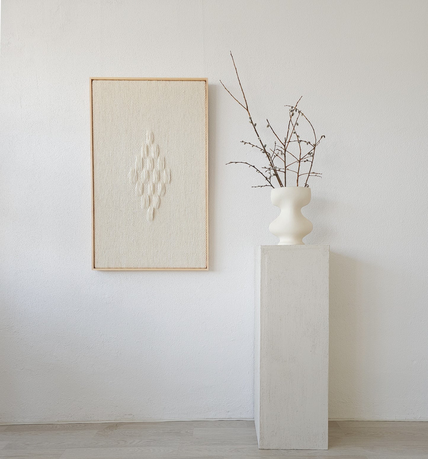 Framed handwoven tapestry featuring a diamond-shaped textured pattern in neutral tones, displayed next to a minimalist white vase on a pedestal, perfect for modern interiors.