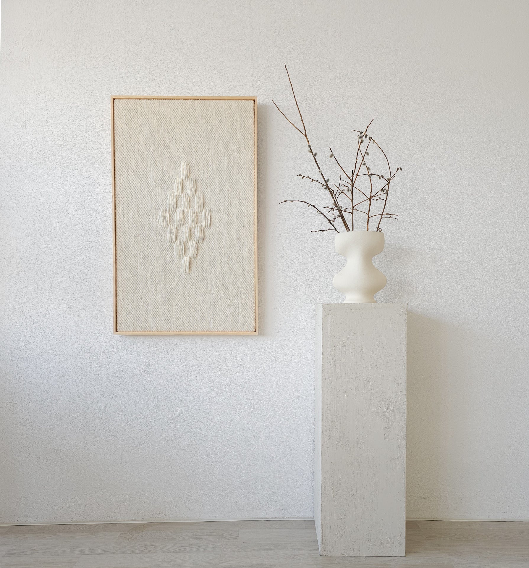 Framed handwoven tapestry featuring a diamond-shaped textured pattern in neutral tones, displayed next to a minimalist white vase on a pedestal, perfect for modern interiors.