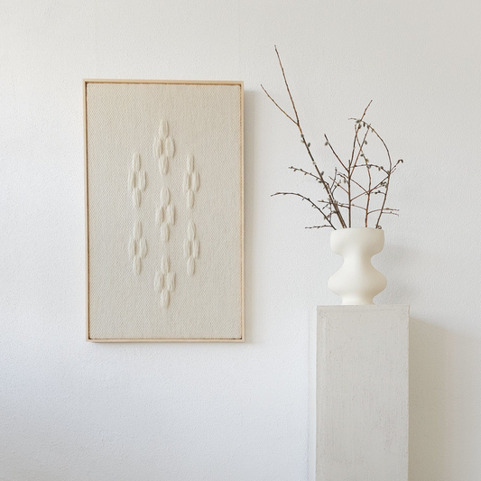 Framed handwoven tapestry featuring a geometric cluster of textured patterns in neutral tones, displayed next to a minimalist white vase on a pedestal, ideal for modern interiors.