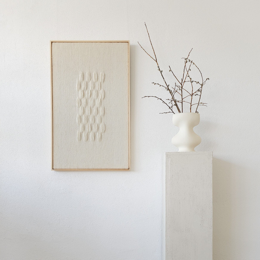 Framed handwoven tapestry in neutral beige tones with a textured pattern, displayed next to a minimalist white vase on a pedestal, perfect for modern interiors.