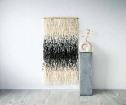 Handwoven shaggy wall hanging in natural cream and black tones, crafted from wool and hung on a bamboo rod, displayed alongside minimalist decor in a modern interior.