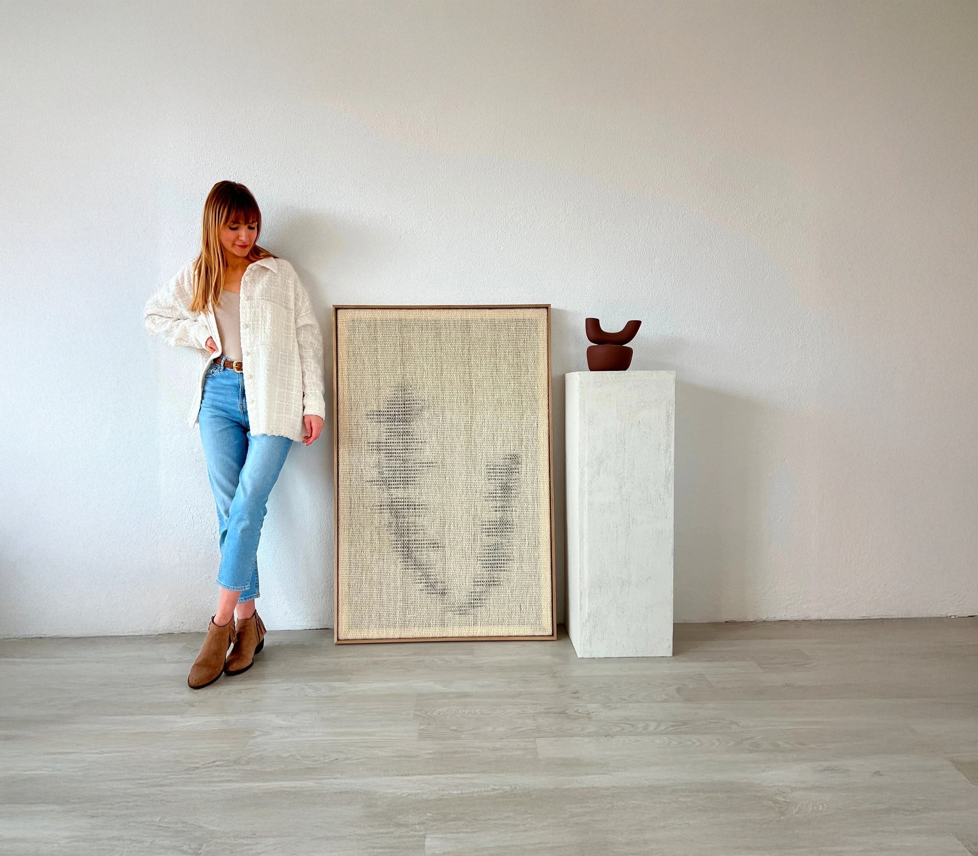 Handwoven wool tapestry in a wooden frame with abstract design, displayed next to a woman for scale and a minimalist pedestal with a ceramic sculpture.