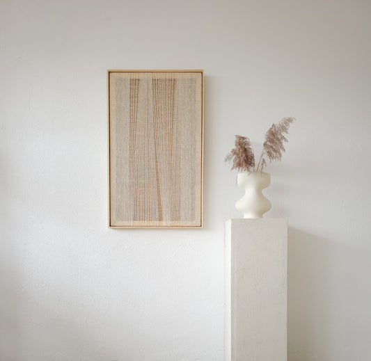 Framed minimalist woven wall art with a natural and textured design in soft beige tones, styled with a decorative white vase holding dried pampas grass on a pedestal.