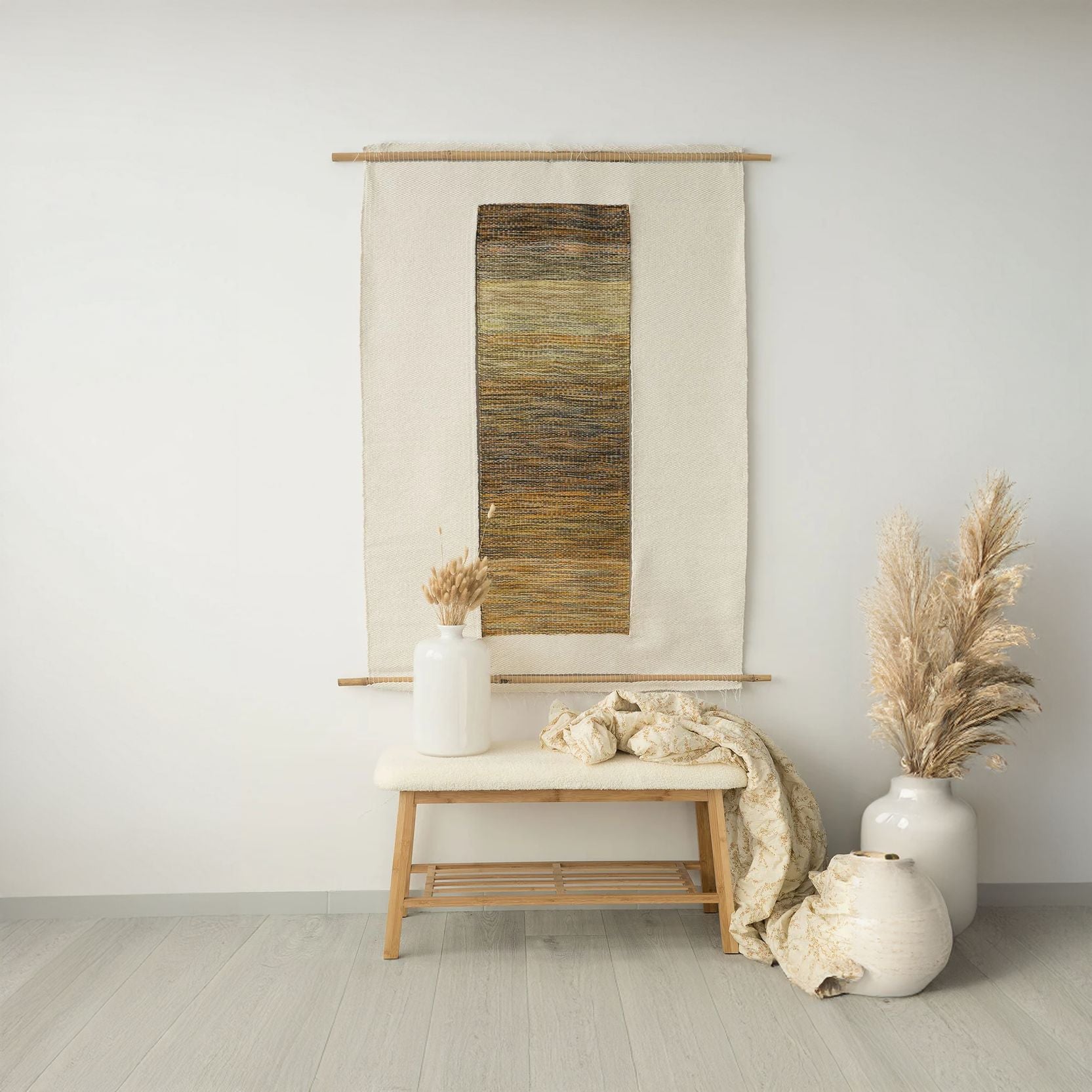 Handwoven wall hanging Kauwela in warm natural tones of yellow, orange, black, and gray, crafted with bamboo and cotton string, styled with a white vase and decorative throw blanket on a wooden bench.