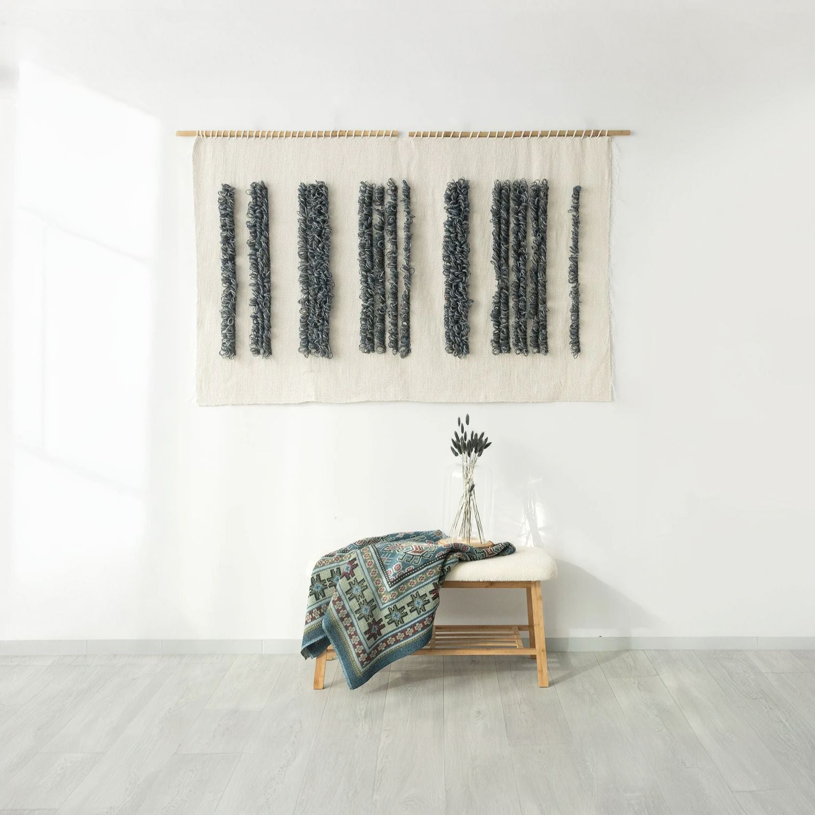 Handwoven Luna wall tapestry featuring a barcode-inspired pattern in natural cotton and shades of blue and gray, suspended on a bamboo rod, styled with a decorative bench and a boho throw blanket, perfect for modern and minimalist interiors.