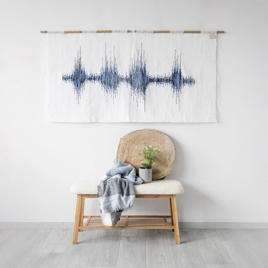 Handwoven white tapestry featuring a blue soundwave-inspired design, suspended on a bamboo rod, ideal for modern and minimalist interiors.