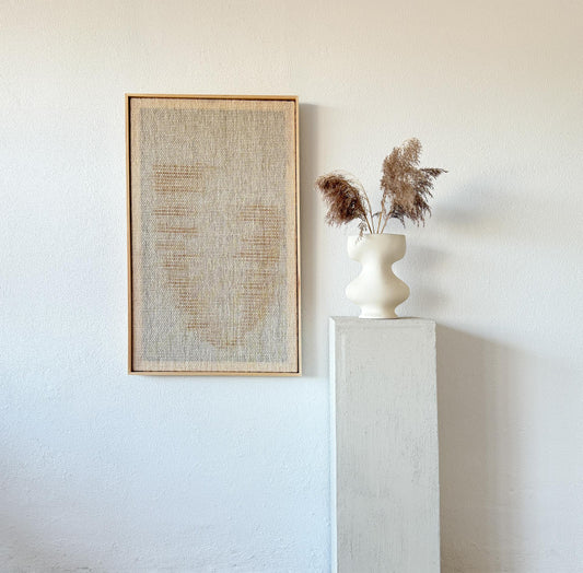 Handwoven wool tapestry in a wooden frame with an abstract yellow design, displayed next to a minimalist pedestal with a ceramic sculpture.