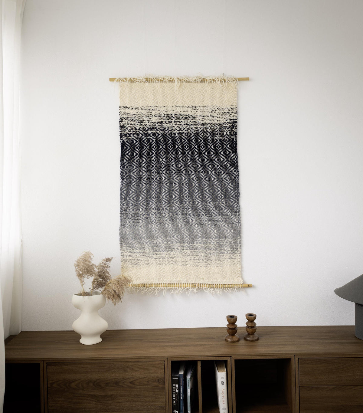BLUO handwoven wool wall hanging with a navy, grey and cream gradient and diamond weave, suspended between two bamboo rods above a wooden sideboard, Lale Studio & Shop