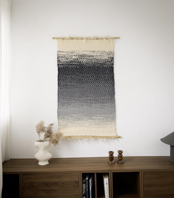 BLUO handwoven wool wall hanging with a navy, grey and cream gradient and diamond weave, suspended between two bamboo rods above a wooden sideboard, Lale Studio & Shop