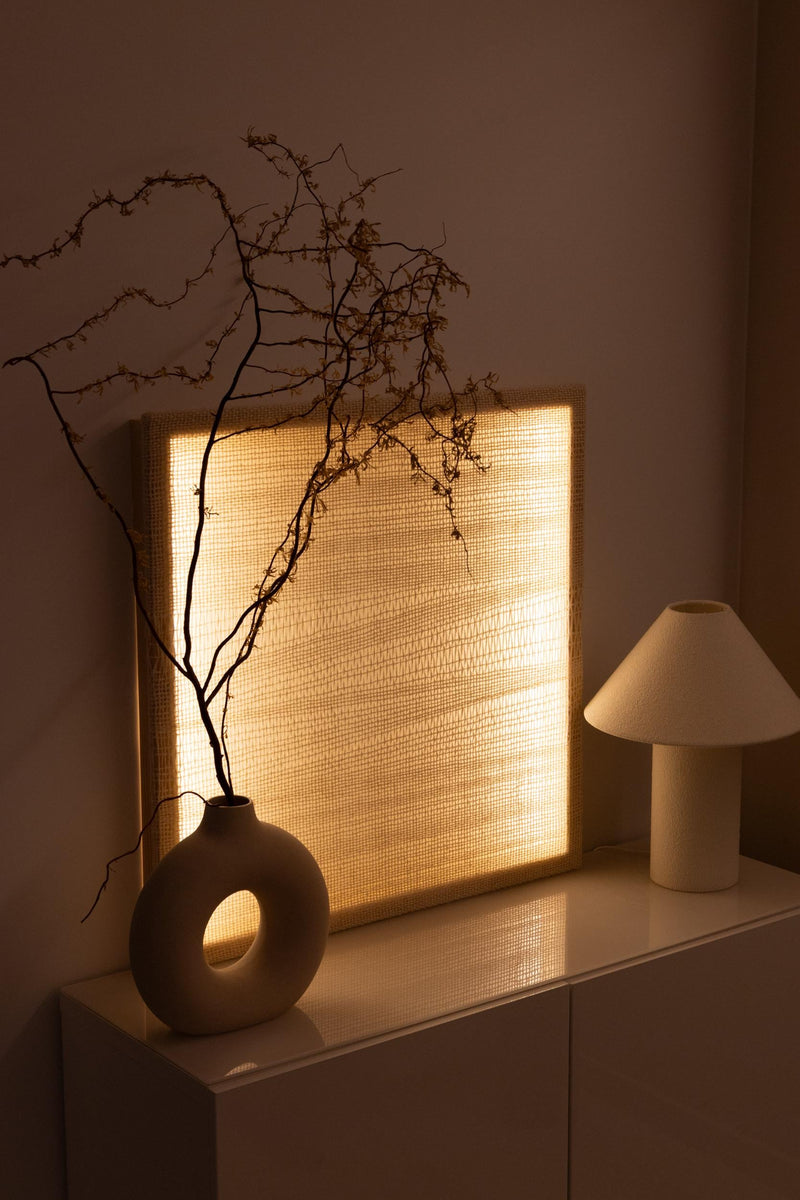 Square illuminated wool tapestry with a translucent handwoven pattern beside a ceramic vase and sculptural table lamp