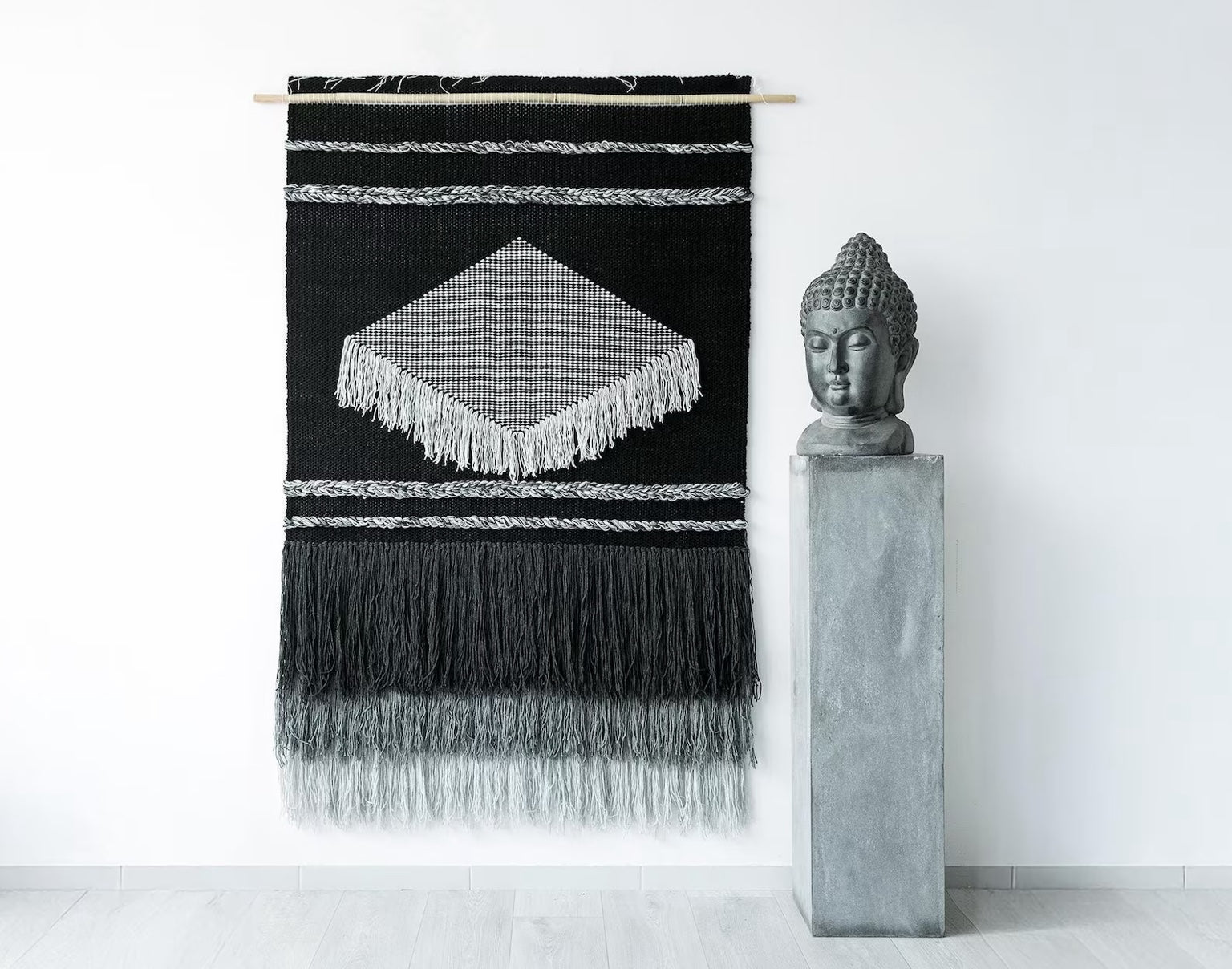 Handwoven black and gray tapestry with intricate geometric patterns, fringed details, and a bamboo rod, perfect for boho and modern interiors.