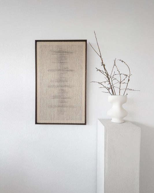 Njolle handwoven neutral beige wall tapestry framed in dark wood, featuring minimalist woven patterns, displayed on a white wall next to a modern vase with delicate branches, ideal for Japandi or Scandinavian interiors.