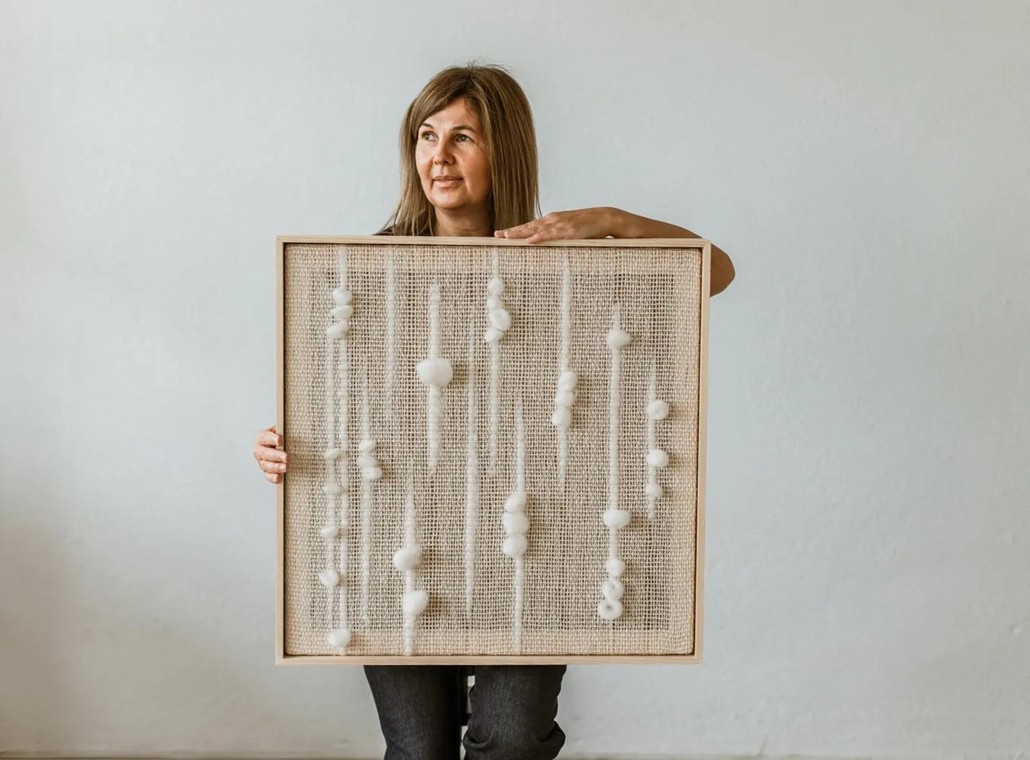 Olit handwoven framed wool tapestry featuring a modern minimalist design with textured white wool patterns, held by a woman in a serene, natural setting. Perfect for enhancing cozy, Scandinavian, or Japandi interiors with unique wall art.