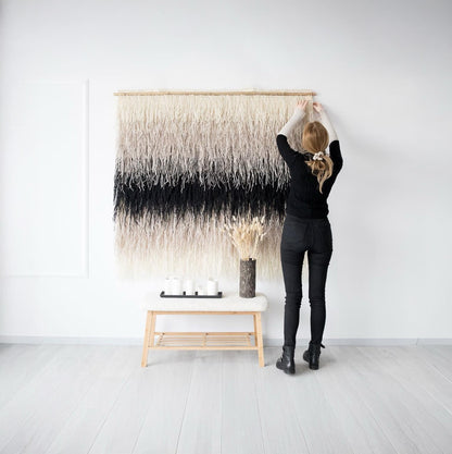 Shaggy Nude handwoven wall hanging in neutral beige and black tones, featuring a layered shaggy texture, being installed on a white wall by a woman. Perfect for adding cozy and modern aesthetics to interiors, from boho to minimalist styles.