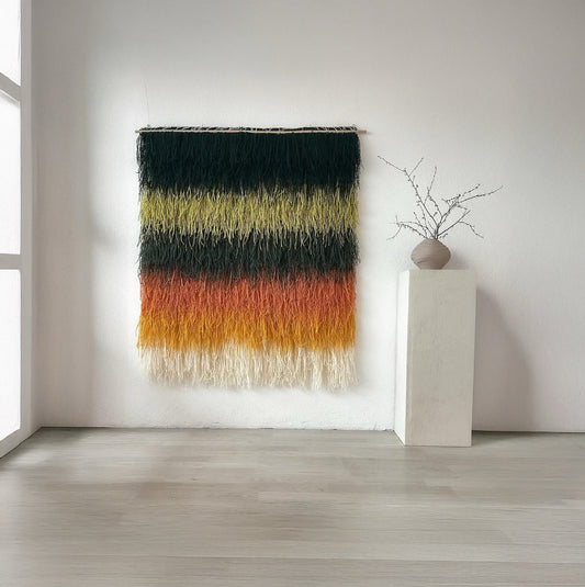 Handwoven wall tapestry with layered fringe design in green, yellow, orange, and beige tones, suspended on a bamboo rod, perfect for boho and eclectic interiors.